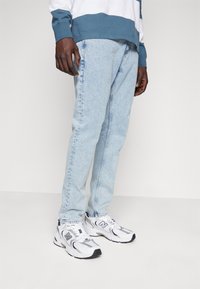Jeans din denim de culoare albastru deschis, cu croială dreaptă, purtați cu adidași albi și gri, având accente negre. Stil casual.