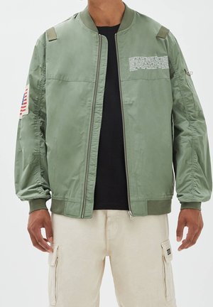 Homme portant une veste bomber vert clair avec le texte NASA sur la poitrine et un patch du drapeau américain sur la manche gauche, pantalon cargo beige.