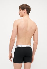 Fekete boxer alsónadrág fehér derékpánttal, rajta a "LACOSTE" logóval. Az anyag simának és rugalmasnak tűnik, így szoros illeszkedést biztosít.