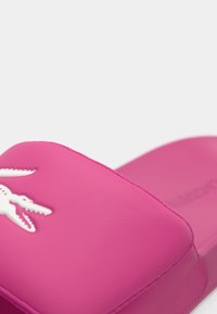 Ροζ σαγιονάρα slide κατασκευασμένη από λείο συνθετικό υλικό, με λευκό λογότυπο κροκόδειλου στο πάνω μέρος και εργονομικό σχεδιασμό πέλματος.