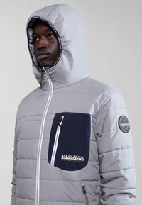 Chaqueta de plumas gris claro con un bolsillo en el pecho de color marino contrastante, cremalleras blancas y un parche con el logo en la manga izquierda. Textura de tejido suave.