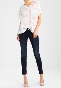 Blouse blanche à motif floral rose avec col en V, associée à un jean skinny bleu foncé et des chaussures rose clair. Tissu fluide, coupe décontractée.