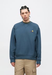 Tmavě modrá mikina s kulatým výstřihem s malým oranžovo-žlutým logem na levé straně hrudi, s žebrovaným límcem a manžetami, v hladké textuře látky.