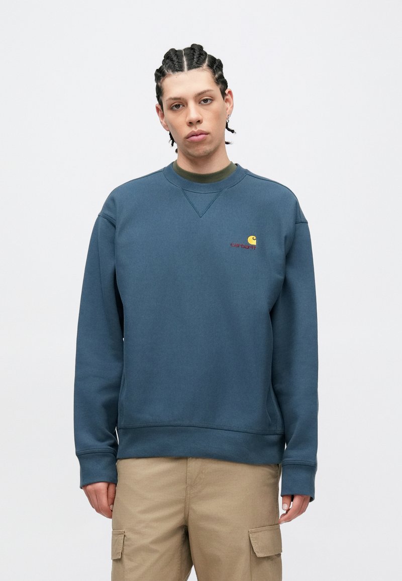 Mørkeblå crewneck-sweatshirt med en lille orange og gul logo på venstre bryst, med ribbet hals og ærmekanter, i en glat stofstruktur.