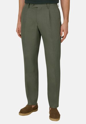 Homme portant un pantalon ajusté vert olive et des espadrilles marron en daim à enfiler, debout devant un fond uni.