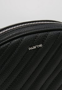 Sac à main noir en similicuir matelassé avec garnitures argentées et un logo proéminent. Présente des coutures diagonales et une fermeture éclair.