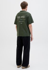 Grøn bomulds t-shirt med korte ærmer, med hvid tekst på ryggen. Sammen med sorte bukser og beige sko, ensfarvet baggrund.