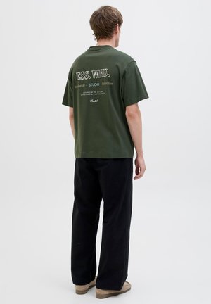 Grøn bomulds t-shirt med korte ærmer, med hvid tekst på ryggen. Sammen med sorte bukser og beige sko, ensfarvet baggrund.