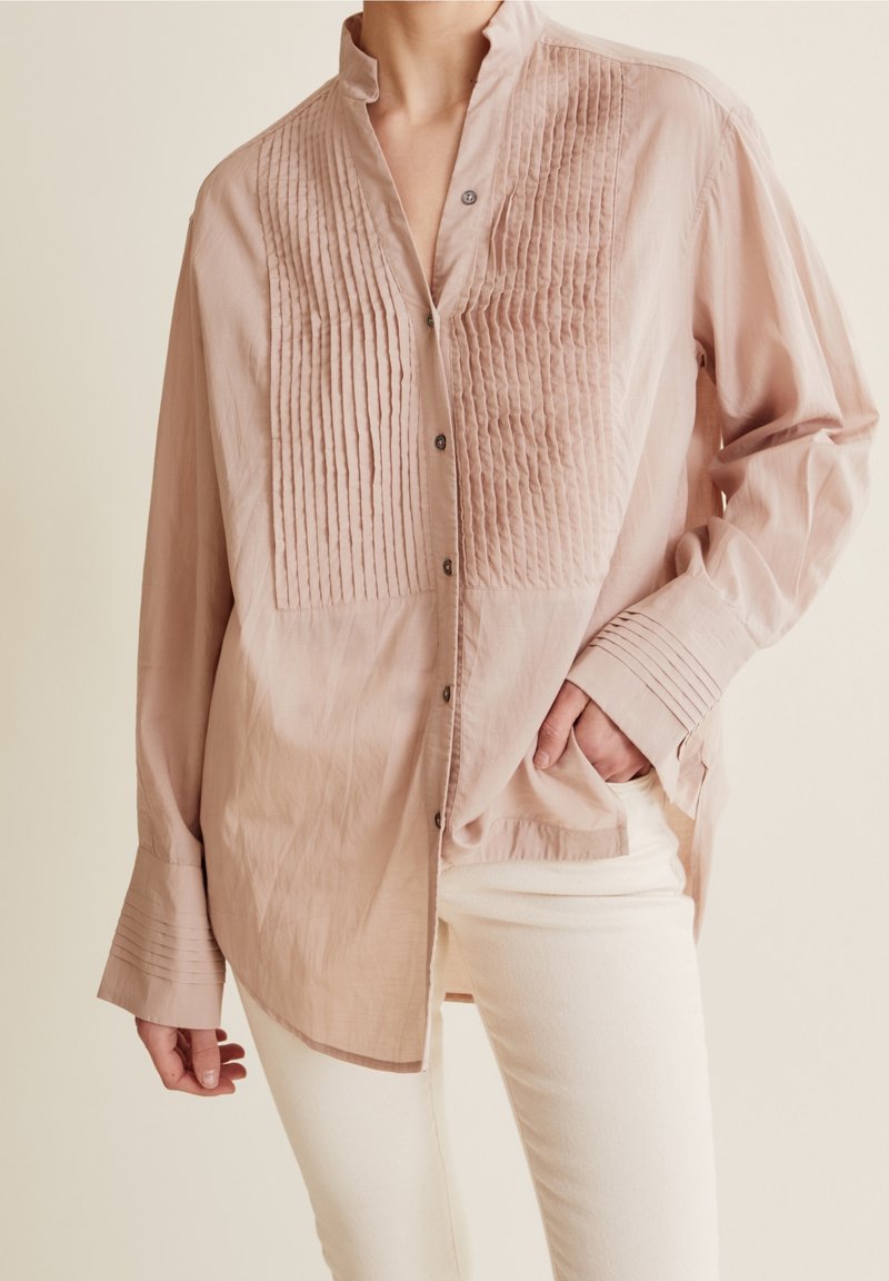 Femme portant un chemisier ample rose clair avec des plis verticaux à l'avant et aux poignets, associé à un pantalon blanc, main dans la poche.