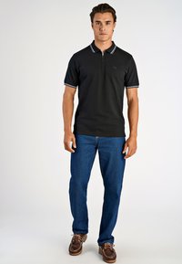 Svart polo tröja med dragkedja i kragen, vita kantband på ärmarna, parat med blå jeans och bruna loafers. Slät tygtextur.