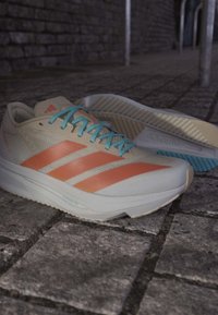 White and orange running shoes with blue laces resting on a cobblestone path under dim lighting.