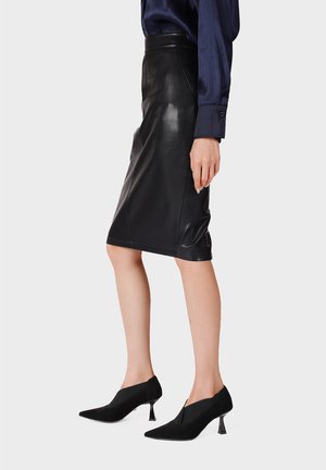 Donna che indossa una gonna nera in pelle alla lunghezza del ginocchio, una camicetta blu scuro a maniche lunghe e scarpe nere con tacco e punta appuntita, in piedi davanti a uno sfondo semplice.