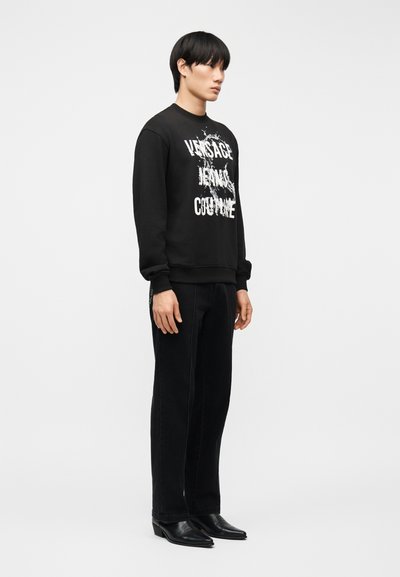 Jeune homme portant un sweat-shirt noir à manches longues avec un texte blanc, un pantalon droit noir, et des chaussures noires pointues, debout de profil.