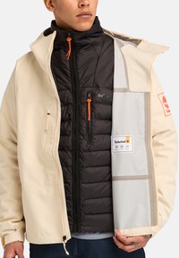 Miesten Timberland-takki on saanut vaaleanbeigen päällisen, mustan eristetyn sisäkerroksen, oransseja yksityiskohtia, vetoketjullisia taskuja sekä näkyvän merkkilapun.