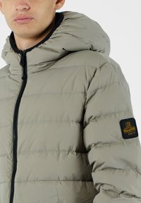 Giacca beige imbottita con cappuccio, dotata di una cerniera frontale e di un accento nero sulla manica con un logo. Il tessuto ha una superficie liscia.