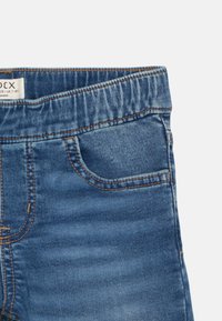Blaue Jeans aus Denim mit elastischem Bund und kleiner Fronttasche, die detaillierte braune Nähte und Stofftextur zeigt.