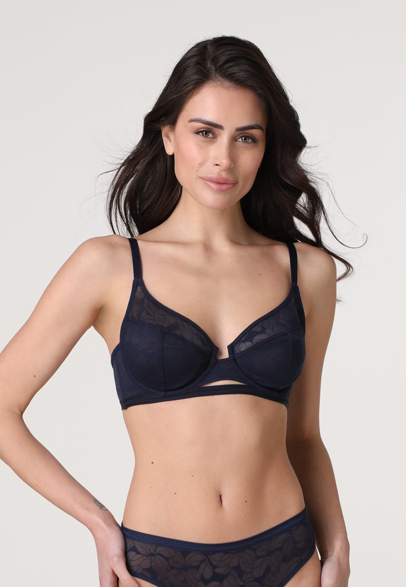 Reggiseno blu navy con coppe in pizzo floreale trasparente e ferretto, dotato di spalline regolabili e una fascia con tagli per un ulteriore dettaglio di design.