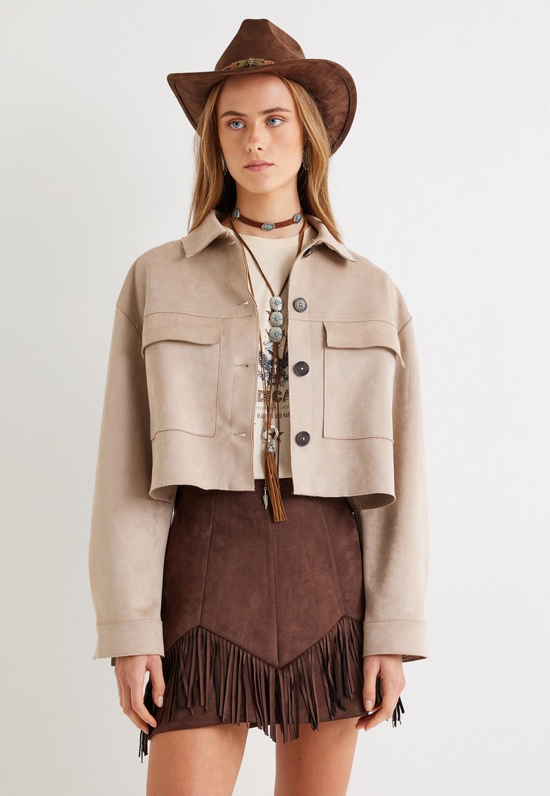 Giacca in suede beige tagliata, con due tasche sul petto, abbinata a una gonna di frange marrone, accessoriate con un cappello marrone e una collana a strati.