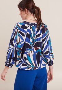 Mehrfarbige Bluse mit kräftigen blauen, schwarzen und braunen Blattmustern auf einem weißen Hintergrund, mit weit ausgestellten Ärmeln und einem Knopf-Detail am Ausschnitt.
