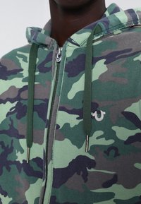 Sudadera con capucha de camuflaje verde, con cierre frontal, cordones ribeteados y un pequeño logo plateado en el pecho.