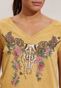 Camisa amarilla de escote en V con un gráfico de un cráneo de toro ornado, decorado con flores rosas, hojas verdes y mariposas alrededor de los cuernos.