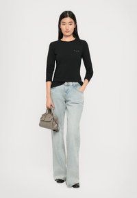 Frau mit langen schwarzen Haaren, die ein schwarzes Langarmshirt, hellblaue weite Jeans, schwarze Schuhe trägt und eine graue Handtasche hält, steht.