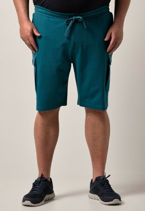 Mand iført turkis cargo shorts til knæet og sorte sportssko står mod en ensfarvet baggrund.