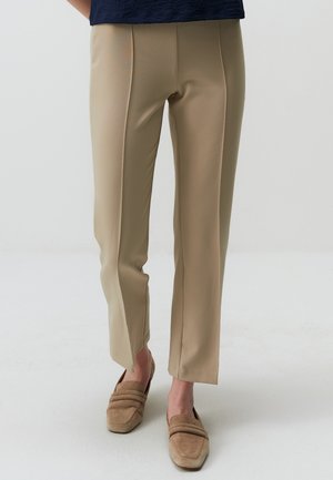 Pantaloni beige su misura con una texture liscia, frontale piatto e sottili spacchi laterali all'orlo, abbinati a scarpe slip-on beige chiaro.