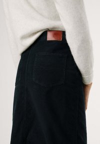 Schwarzer Cordrock mit einer einzelnen Gesäßtasche und einem braunen Lederlogo-Patch in der Nähe der Taille. Ein hellgrauer Pullover ist teilweise sichtbar.