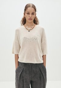 Beige gestricktes Oberteil mit lockerer Passform, kurzen Ärmeln und einem strukturierten Muster entlang des Ausschnitts, kombiniert mit dunkelgrauen, gefalteten Hosen.