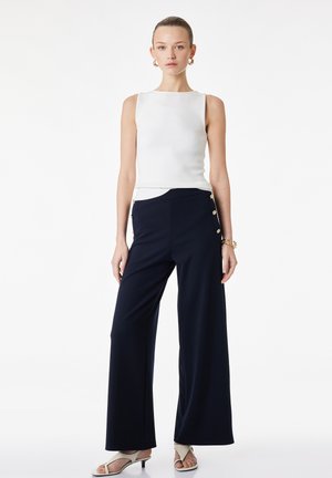 Vrouw draagt een mouwloze witte top, marineblauwe wijde broek met hoge taille en zijknopen, witte sandalen met hak en gouden oorringen en armband.