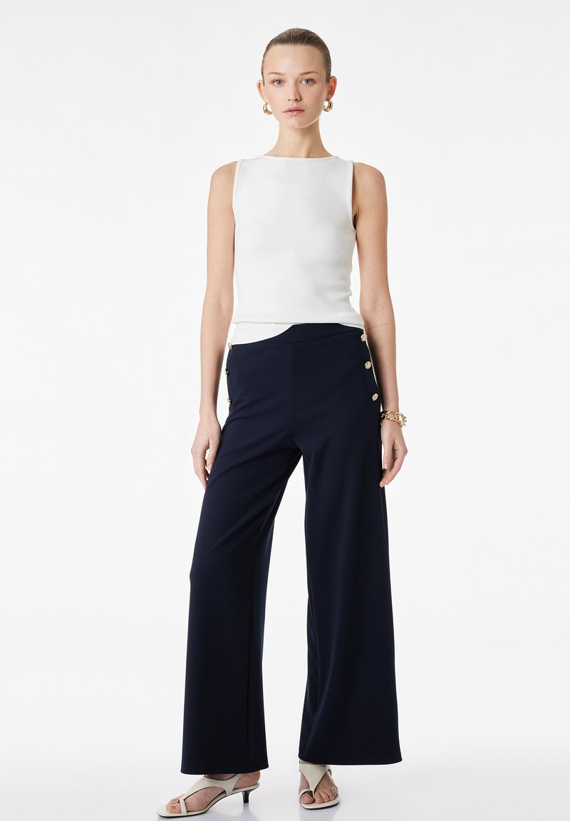 Femme portant un haut blanc sans manches, un pantalon large taille haute bleu marine avec des boutons sur le côté, des sandales blanches à talons, et des boucles d'oreilles créoles dorées ainsi qu'un bracelet.