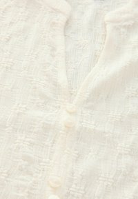 Cardigan blanc, texture tricotée, avec un motif subtil. Cinq petits boutons ronds, semblables à des perles, le long de l'encolure avant. Tissu doux.