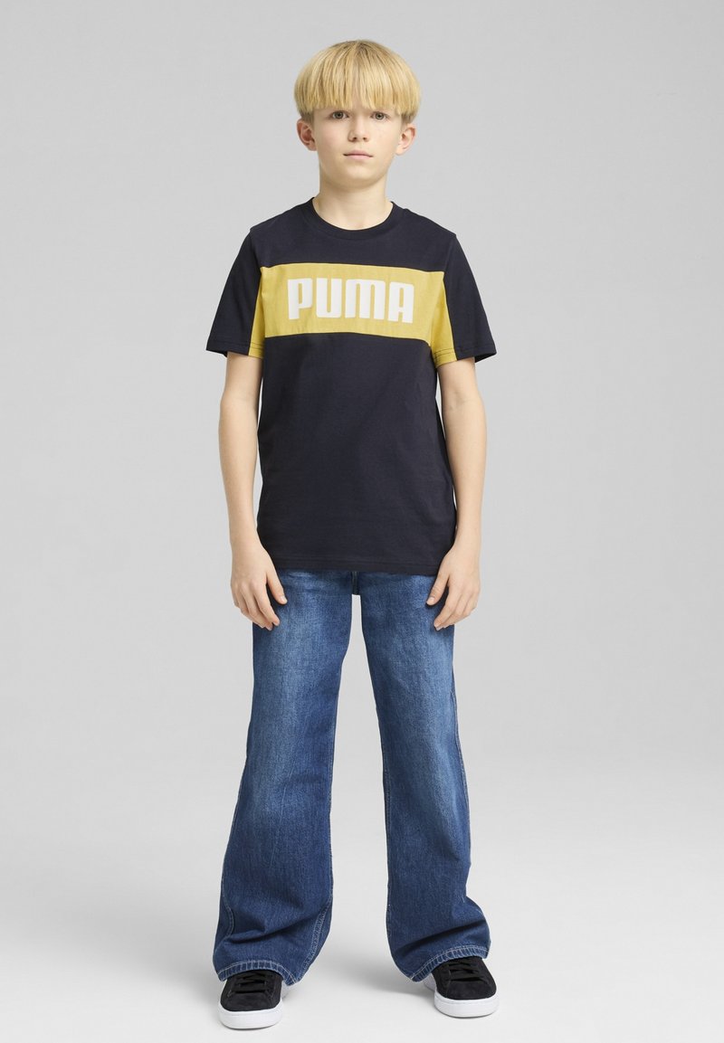 Zwarte en gele T-shirt met korte mouwen en het "PUMA" logo, gecombineerd met blauwe wijde jeans en zwarte schoenen, staande tegen een grijze achtergrond.