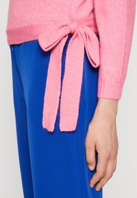 Osoba v modrých kalhotách a růžovém sv sweateru uvázaném na pase mašlí, pravá ruka volně po boku.