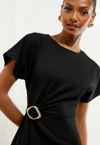 Schwarzes Kleid mit kurzen Ärmeln, das ein verdrehtes Design in der Taille aufweist und eine goldene, unregelmäßig geformte Schnalle als Akzentdetail hat.