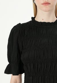 Svart topp med rynket tekstur, korte pufferm, og en volangkant ved halsen. Stoffet ser lett og noe krøllete ut.