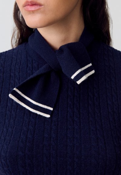 Suéter de punto color navy con un patrón de trenzas texturizado; presenta un cuello atado con acentos de rayas blancas, ofreciendo un aspecto estructurado y sofisticado.