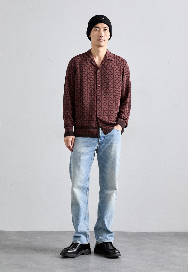 Camicia button-up color bordeaux con un motivo geometrico, abbinata a jeans dritti blu chiaro e stivali in pelle nera. Berretto di maglia nero.