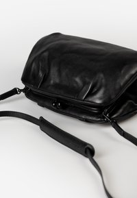 Borsa a tracolla nera in pelle con una superficie liscia, pattina pieghevole e tracolla regolabile. Presenta un branding discreto vicino alla cerniera.