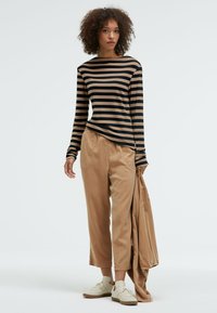 Langärmliges gestreiftes Oberteil in Beige und Schwarz, kombiniert mit beigen weiten Hosen. Die Schuhe sind hellbeige; sie hält eine passende beige Jacke.