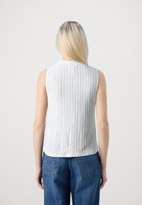 Una donna con lunghi capelli biondi, che indossa un top bianco senza maniche in pizzo e jeans blu, in piedi con le spalle rivolte alla macchina fotografica.