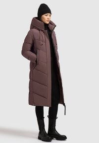 Lang polstret frakke i mauve med høj krave, lynlås fortil og sidelommer. Tykt stof med en quiltet mønster, parret med sorte støvler.