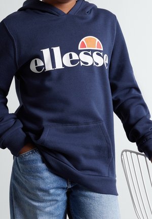 Person trägt einen dunkelblauen ellesse-Hoodie mit Logo, hat die Hände in der Vorderhosentasche und trägt hellblaue Jeans, steht neben einem weißen Stuhl.