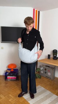 Svijetlosivi sling ruksak od tkanine s okruglim oblikom, s podesivim remenom nošenim preko tijela. Model nosi crnu majicu i plave traperice.