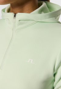 Primo piano di una persona che indossa una felpa con zip verde chiaro, dotata di colletto alto e un piccolo logo bianco sul petto.