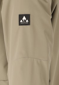 Beige jas met een gladde textuur, voorzien van een zwart vierkant logo-embleem met het opschrift "WHISTLER" op de linker mouw. Eenvoudig ontwerp.