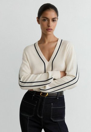 Kvinde med mørkt hår i en hvid V-hals sweater med sorte striber, krydser arme, iført mørke jeans med hvide syninger og sort bælte.