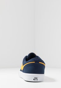 Laivastonsininen kangaskenkä, jossa on keltainen logo, pyöristetty kanta, valkoinen kumipohja ja teksturoitu päällinen, jossa on hienovaraisia ompeluja ja brändielementtejä.