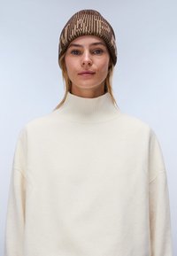 Kvinde iført en ribbet beige rullekravetrøje og en brun mønstret strikket hue mod en ensfarvet lys baggrund.